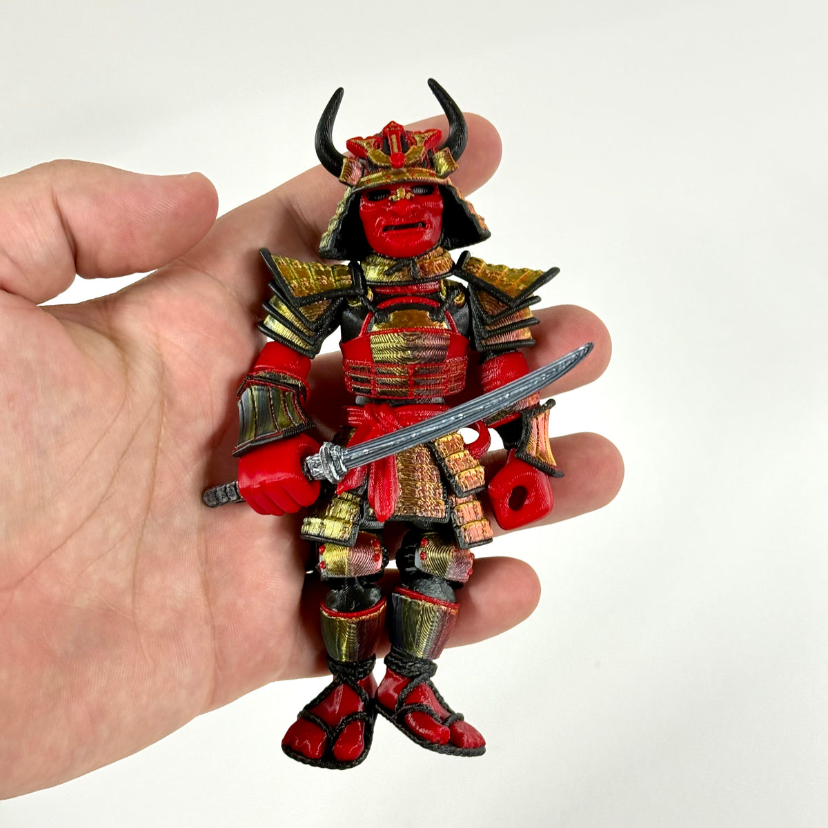 A red and gold samurai warrior action figure held in a person's hand against a plain white background.