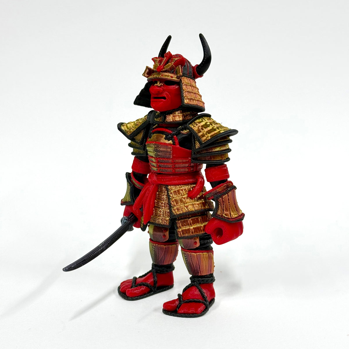A stylized samurai warrior action figure or collectible statue, featuring red skin and armor with gold and black details, holding a curved sword, photographed against a plain white studio background.