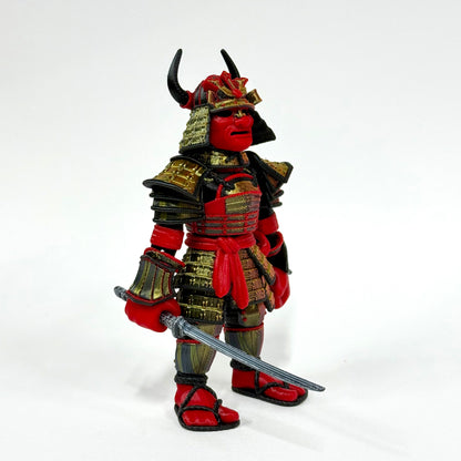 A red and black samurai action figure with gold armor plates, a horned helmet, and a silver sword, photographed against a plain white studio background.